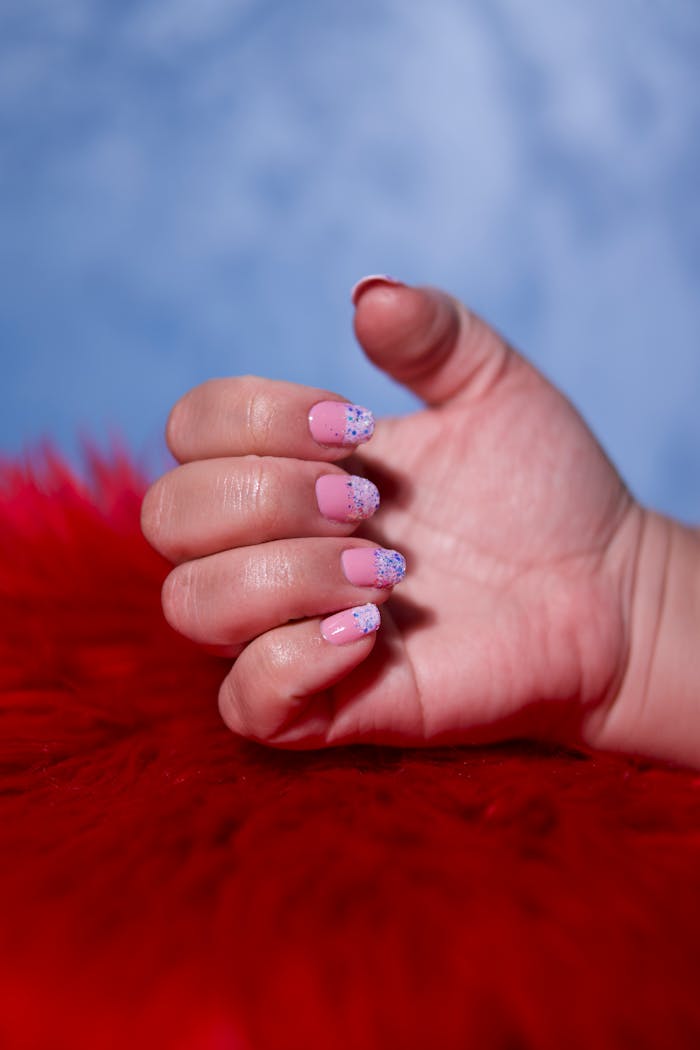 Close-up of a hand with pink nails and glitter design on red fluffy surface.