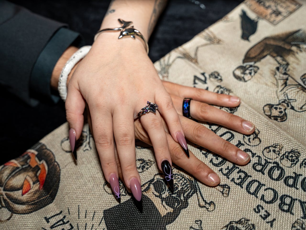 about-img Close-up of hands with Halloween nail art and jewelry on themed fabric background.