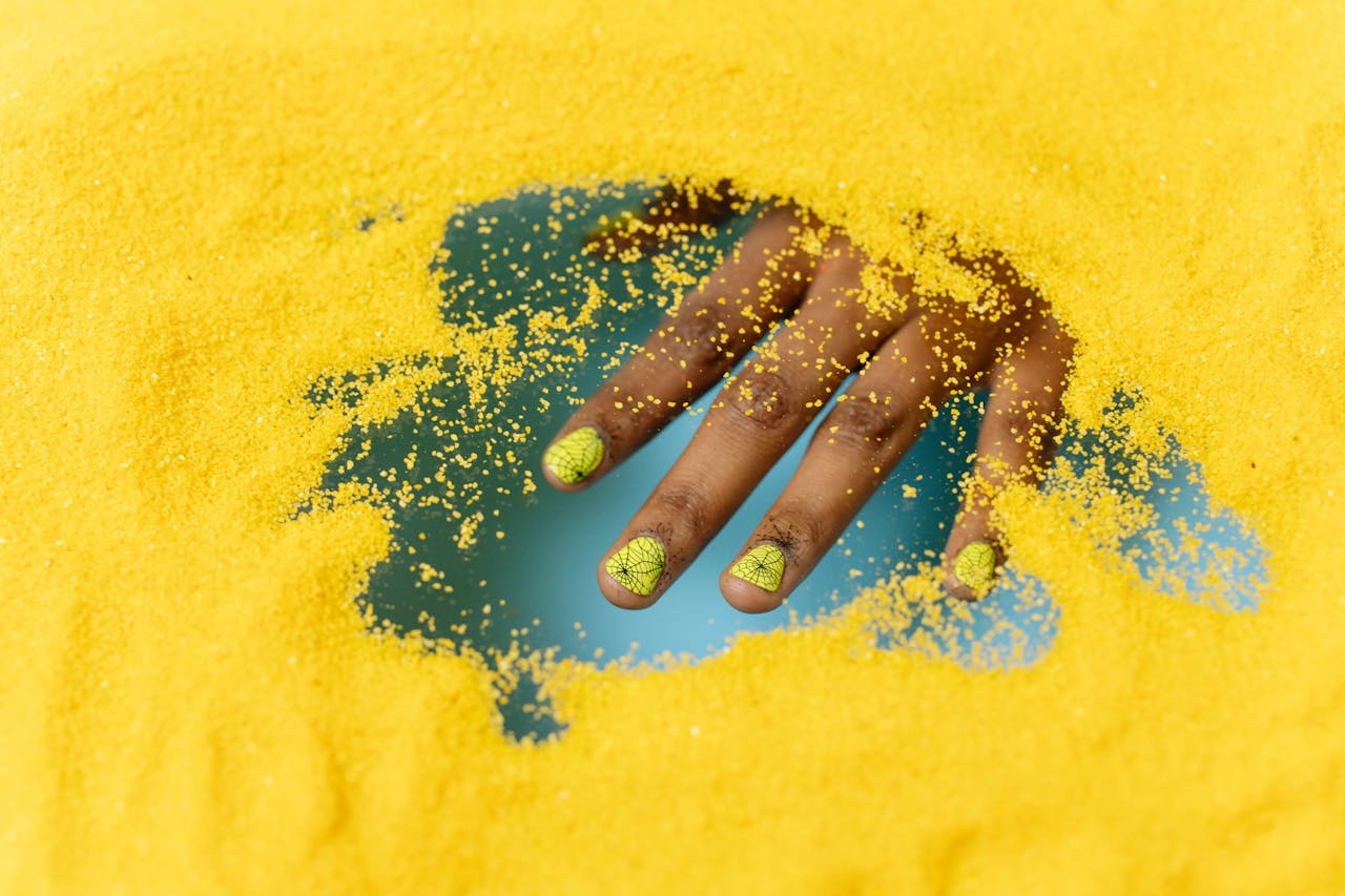 hero-img-01 Close-up of a hand with creative nail art partially buried in vibrant yellow sand.