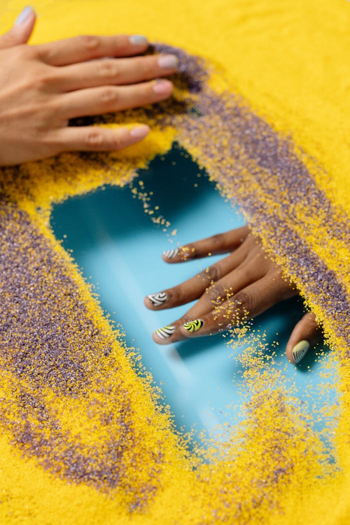 gallery-6 Close-up of hands with intricate nail art on colorful sand, featuring vibrant designs and reflections.